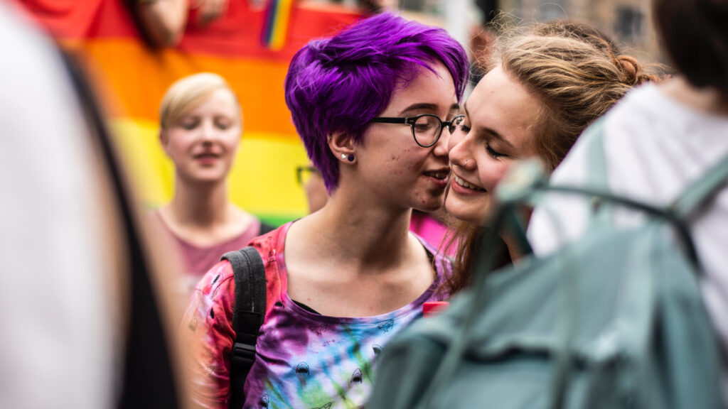 Frau mit rot-weißem Hemd vor einer Regenbogenfahne