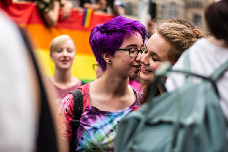 Frau mit rot-weißem Hemd vor einer Regenbogenfahne